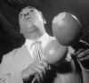 A dark-skinned man wearing a light-colored suit and tie and holding two maracas is seen from below at an angle.