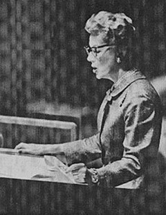 Monochrome photograph of a bespectacled, short-haired woman in a suit jacket reading from papers at a podium