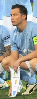 A man with short dark hair kneels on a football pitch before a match, wearing a light blue shirt, white shorts and light blue socks. He is holding a pennant.