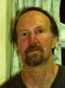 A middle-aged man with light brown hair and a goatee stands in front of a window