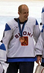 A Caucasian ice hockey player dressed in a blue jersey with green and white trim, as well as a blue helmet. He stands relaxed on the ice, holding his hockey stick in one hand.