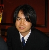 A 39-year-old Japanese man wearing a black suit coat, a blue tie, and a white button-up shirt. He has black hair and brown eyes, and is against a dark red background.