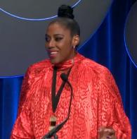 Broom stands behind a podium and speaks into the microphone. She wears a peach silk dress. She has brown skin and her hair is styled in a bun on top of her head.