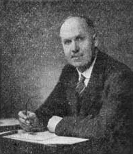 A black-and-white photograph of a man in a suit sitting at a desk, holding a pen, and looking towards the camera.