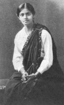 A young Indian woman wearing a dark sari over a white blouse; seated, hands folded in her lap. Her hair is parted center and dressed back behind her ears.