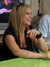 A blonde-haired woman in a black shirt smiles while sitting at a green table.