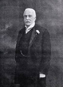 A vintage photograph of an elderly man with white hair and a moustache, wearing a dark suit with a white shirt, a bow tie, and a flower on his lapel, standing and looking slightly to the side.