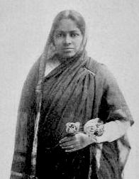 An Indian woman wearing a dark sari with the tail draped over her head and shoulder. She is holding flowers.