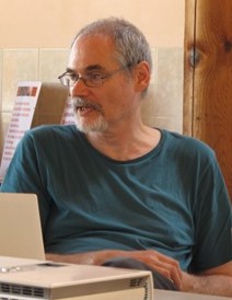 A man with grey hair and glasses, wearing a teal shirt, sitting and looking to the side with a relaxed posture.