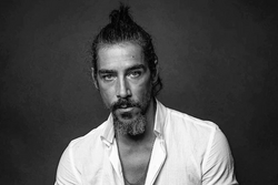 black-and-white image of Óscar Jaenada with goatee and top bun, wearing an open-necked white shirt and staring intently at camera