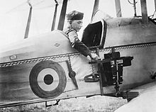 a military member is seated in the observer's cockpit of a Royal Flying Corps plane. The emblem of the Royal Flying Corps is prominently displayed on the side of the aircraft. The military member is seen adjusting a camera mounted to the side of the fuselage, positioned just behind the wing.