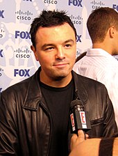 A man with black hair, wearing a leather jacket, and being interviewed. There is a small microphone in front of him, with a television channel logo placed on it.