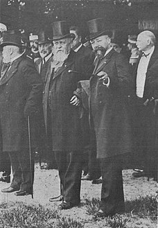 Photo en noir et blanc d'hommes portant pour la plupart des chapeaux ; au premier plan, deux hommes échangent : celui à gauche a une barbe blanche et celui de droite, plus mince, arbore une barbe plus foncée mais grisonnante