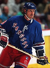 A hockey player stares intently to his left. He is in full uniform with a blue helmet and jersey, and red pants. The jersey has the word "RANGERS" spelled diagonally down his chest.