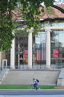 Une photo du John Glenn College of Public Affairs, avec un drapeau américain suspendu à l'intérieur et un cycliste passant devant les marches en pierre.