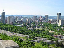 A tree-lined highway is in the foreground, angled diagonally from bottom right to middle left of the image. Buildings are in the centre, and the background is a sky meeting rolling hills in the distance.