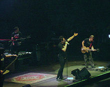 Four people. On the far left a man with a low hand. Mounted on a platform a man sits on a bench with a keyboard in front. In the center a man standing, wearing black with his left hand and taking a microphone with his right hand extended. On the far right a man semi-inclined holding a guitar.