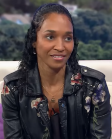 A black woman with wavy black hair. At her hairline, her hair is styled into a smooth swoop