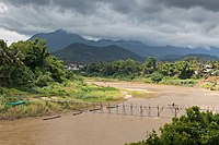 Landscape with a wooden footbridge crossing the Nam Khan river, where two workers are working at the consolidation of this structure, holding a big beam during the monsoon