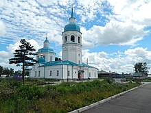 Photographie d'un bâtiment religieux orthodoxe blanc blanc avec un clocher au toit bleu et une chapelle à l'arrière possédant une coupole bleue, le tout au milieu d'un espace vert.