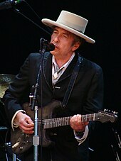 A man behind a microphone stand, wearing a white hat and black suit. The man is playing the guitar and singing into the microphone.