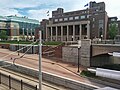 Washington Avenue's light rail tracks, pavement, and bus stop, in front of a retaining wall, on top of which sits Coffman's front lawn, with Coffman itself directly behind it. Slightly to the left of Coffman is the glass facade of Nils Hasselmo Hall.