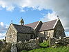 A stone church with slate roofs seen from the southeast. To the right is the chancel, to the left is a large transept, beyond which can be seen a bellcote