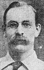 Black and white photo of a middle-aged man wearing eyeglasses, a handlebar moustache, white-collar dress shirt and a necktie