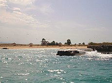 Picture of a sandy shore delineated from both sides by dark brown rocky formations. The sandy shore is at the edge of a wavy blue colored body of water. At the top the light blue sky shows some white clouds and some sea birds.