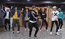 Seven young men performing synchronised dance moves, wearing casual clothing. Some of them have dyed hair.