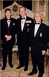 A color photograph of three men standing together, each wearing black jackets and black trousers. The jacket is open in a deep V-shape toward the waist where it buttons. Each man is wearing a bow tie over the collar of their white shirts and each has a pocket handkerchief sticking out of their breast pocket. Their shoes are black.