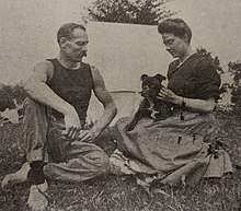Black and white photo of a middle-aged man and woman wearing casual clothes, sitting outside of a tent, playing with a small dog