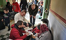 People sitting on a tiled floor, some eating food wrapped in paper