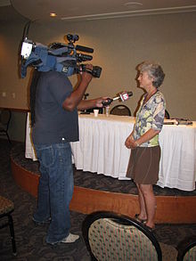A man holding a camera and a microphone with a red mic flag with a white "16" interviews a woman at an event.