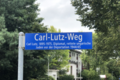 Photographie d'un panneau signalétique indiquant en allemand en lettres blanches sur fond bleu: "Carl-Lutz-Weg" dans une grande typographie et "Carl Lutz, 1895-1975, Diplomat, rettete ungarische Juden vor der Deportation, 1944/45" dans une typographie plus petite ; au loin, des maisons et des arbres au feuillage vert.