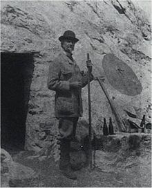 Photographie d'un homme se tenant debout devant l'entrée d'une grotte, un bâton ferré à la main.