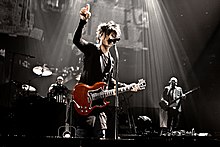 Groupe Indochine sur scène dont Mr chanteur avec une guitare rouge en train de chanter le point le