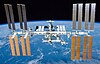 A planform view of the ISS backdropped by the limb of the Earth. In view are the station's four large, gold-coloured solar array wings, two on either side of the station, mounted to a central truss structure. Further along the truss are six large, white radiators, three next to each pair of arrays. In between the solar arrays and radiators is a cluster of pressurised modules arranged in an elongated T shape, also attached to the truss. A set of blue solar arrays are mounted to the module at the aft end of the cluster.