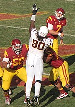 An American football player in a white, gold, and brown uniform reaches to block a pass by another player. He is being blocked by two other players in red and yellow uniforms.