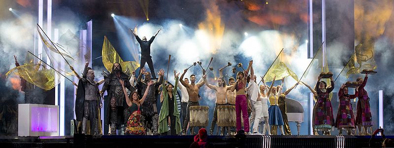 Photograph of performance of "Love Love Peace Peace" at the 2016 final: Petra Mede and Måns Zelmerlöw perform on stage surrounded by performers dressed in costumes of past Eurovision acts