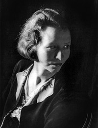 A moody photograph of Eda St. Vincent Millay in which her hawk-like visage is half-hidden in shadow, and she is glancing over her shoulder to the right with a supercilious air.