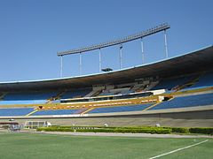 Seção lateral de um estádio oval, com assentos azuis e brancos vazios, vista do campo; o céu está azul e sem nuvens.