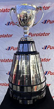 A trophy in the design of a silver cup affixed to a large, round wooden base. The base has silver plates attached to it engraved with the names of previous winners.