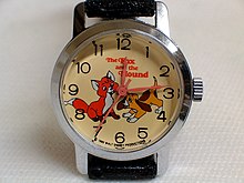 Montre sur présentoir avec un bracelet usagé noir replié derrière, le cadran de la montre est orné d'une image d'un renardeau et un chiot, les personnages du film.