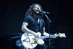 25 year-old Le'aupepe sings into his microphone on a stand while playing his guitar. His eyes are almost closed. He has over shoulder-length dark hair and a beard. He wear a black short-sleve shirt and has tattoos on both arms