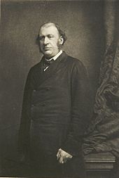 A black-and-white photograph of James Fitzjames Stephen, a balding man with sideburns. He stands looking to the left of the camera near a curtain. He is holding a cane in his left hand.