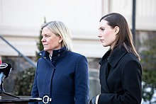 Two women in dark blue coats stand behind a podium looking to the left.
