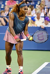 An African American woman in all white clothing, shirt, bandanna, and skirt, is starting the serve of a tennis ball