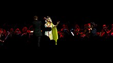 Image d'une chanteuse habillée en blanc et vert avec un masque florale jouant avec un orchestre