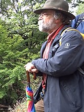 Profil d'un randonneur aux cheveux blancs, portant des lunettes et un chapeau, appuyé sur un parapluie.